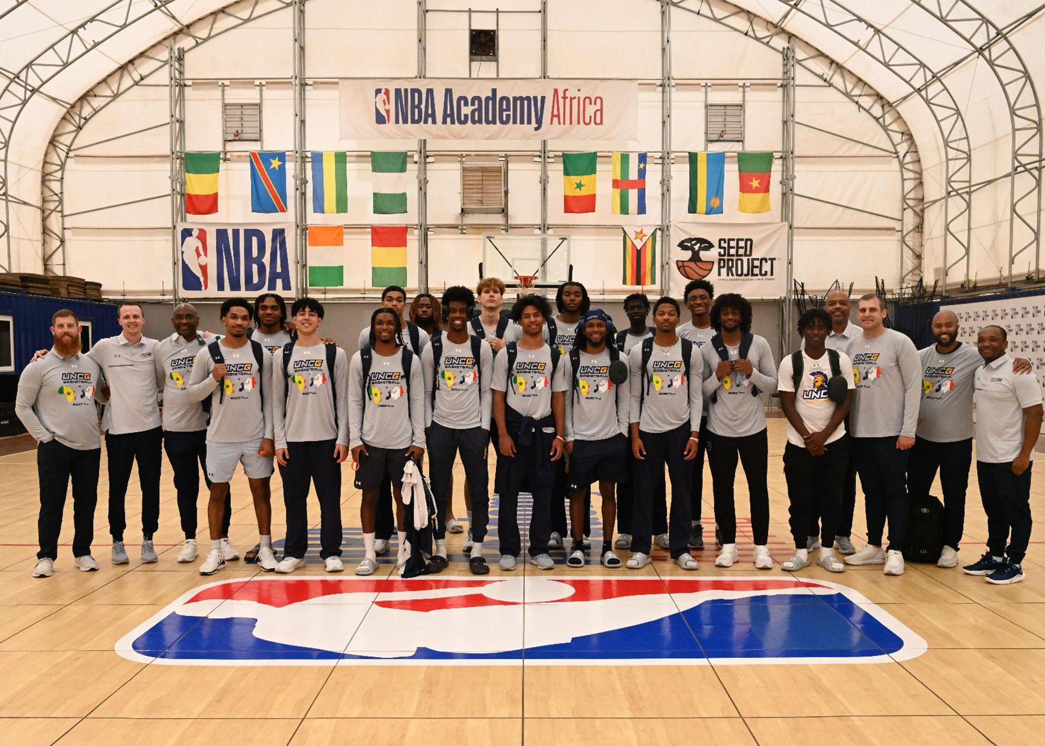 UNCG Men's Basketball team poses together in an arena with international flags and NBA Academy Africa sign behind them. 