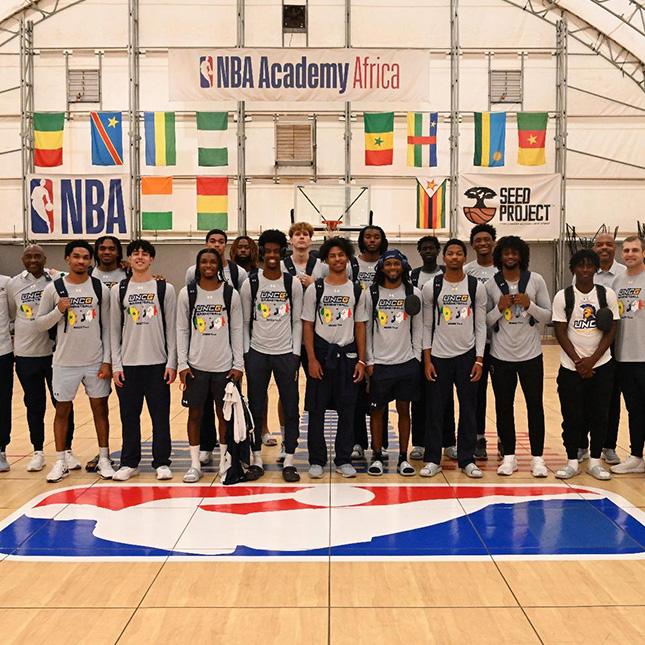 UNCG Men's Basketball team poses together in an arena with international flags and NBA Academy Africa sign behind them.