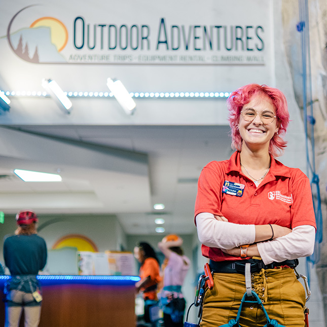 Woman stands in front of the Kaplan Center Outdoor Adventures desk in a climbing wall harness.
