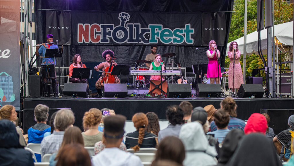 Band plays in front of a NC Folk Fest banner with filled audience seats in the foreground.