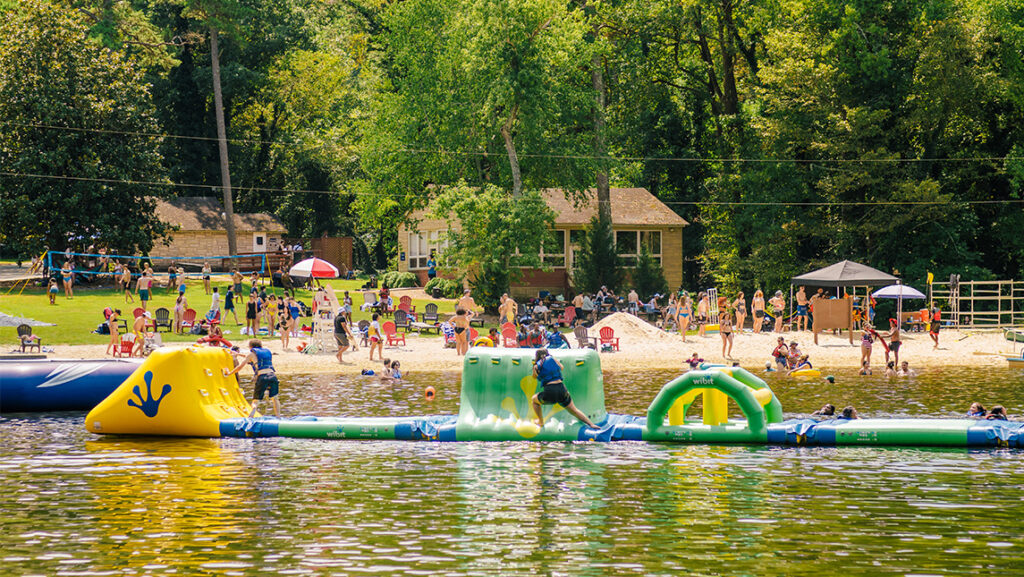 An inflatable obstacle course on a lake with a sandy beach in the background and lots of students in the water and on the beach.