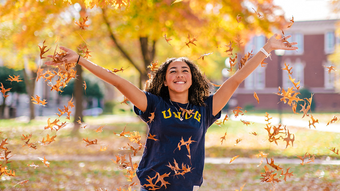 Girl in UNCG t-shirt stands on campus and throws yellow fall leaves up in air.
