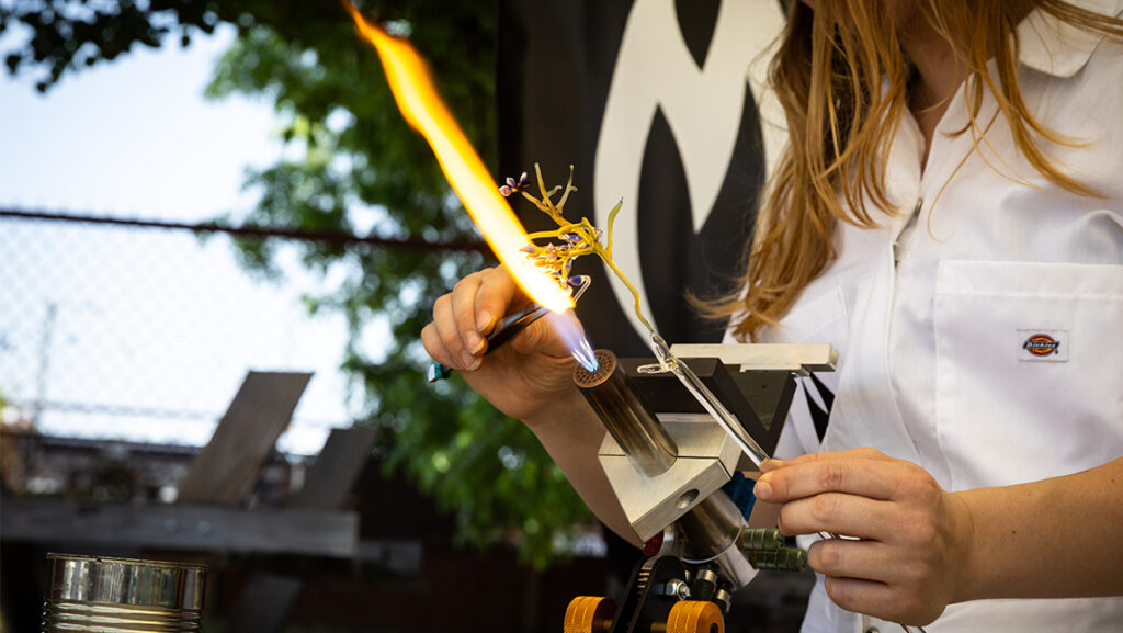 A woman uses a blowtorch to shape glass.