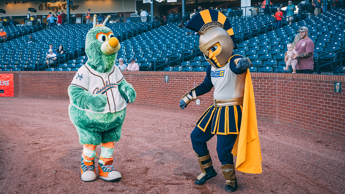 The UNCG mascot Spiro dances on a ballfield with the Greensboro Grasshoppers mascot.