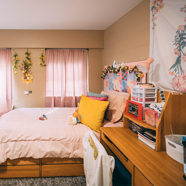 Dorm room decorated with pink and yellow accents and wall hangings. 