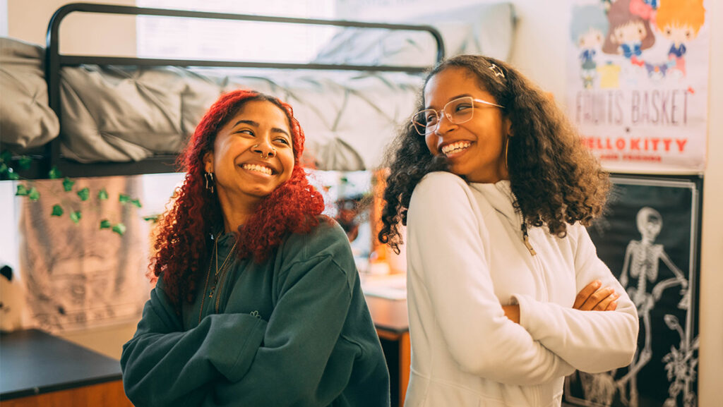 Two women stand back to back with arms crossed in a dorm room as they smile at each other.