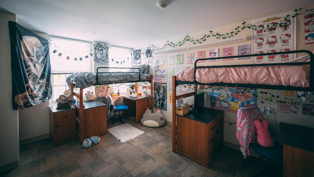Dorm room with loft beds and desks decorated with Hello Kitty wall hangings on one side and skull flag on the other. 
