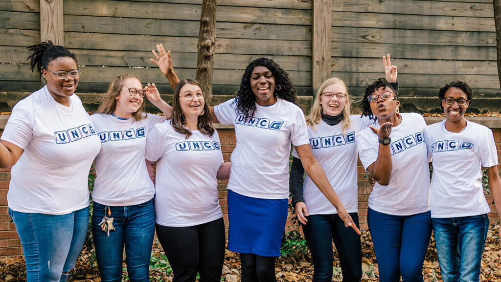 Seven women pose together in t-shirts with UNCG letters that look like periodic table elements.