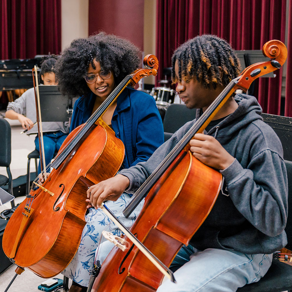 Two students play the cello in a UNCCG rehearsal hall with other musicians.