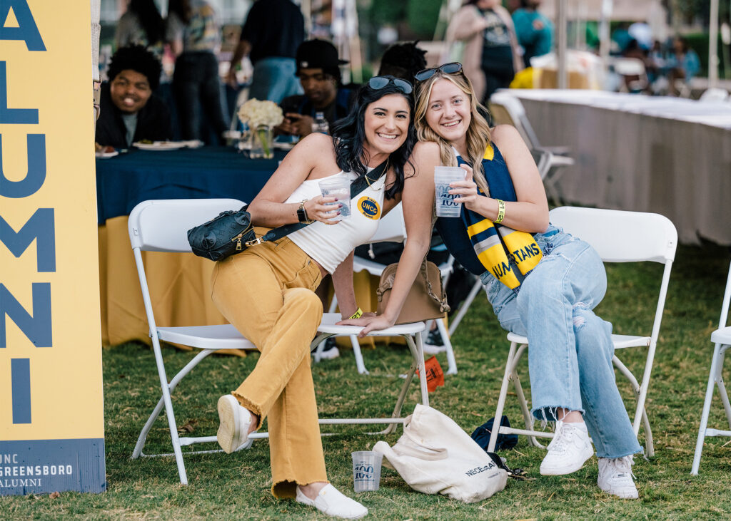 Two women wearing UNCG colors and holding drinks pose in folding chairs with tents and tables behind them.
