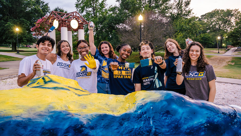 Group of students stand behind a rock they are painting blue and gold with the clock tower in the background. 