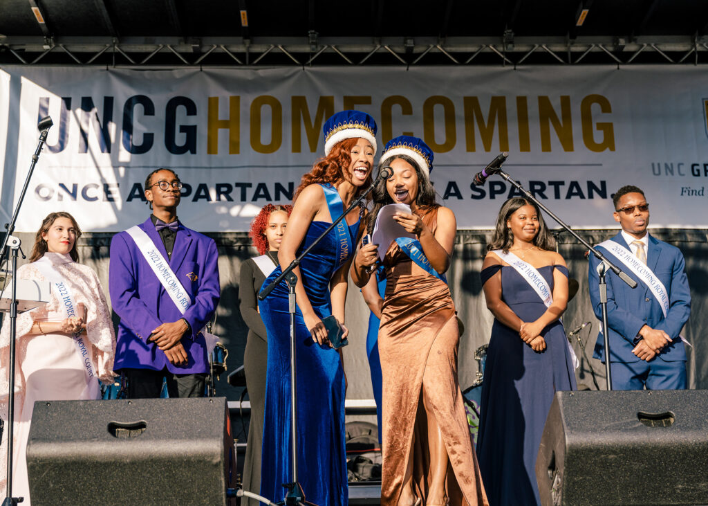 Women and men in formal wear, crowns and sashes stand on a stage with a UNCG Homecoming banner behind them.