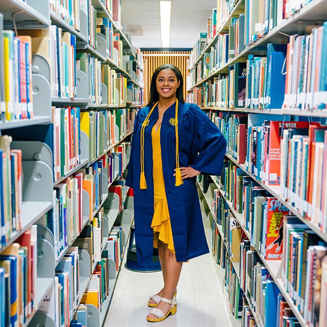 A student in a graduation gown poses in a library aisle.  Student