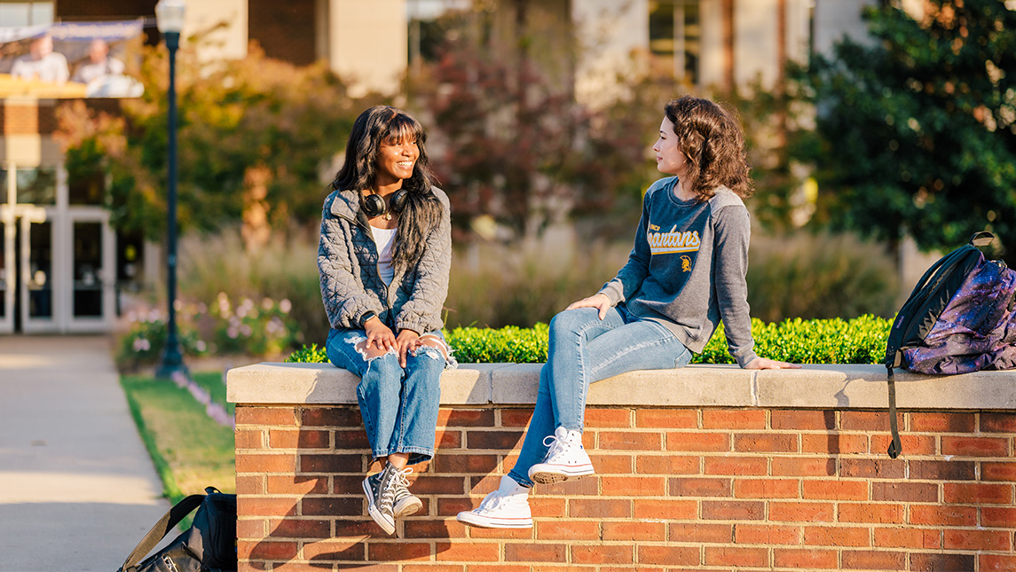 Two women chat as they sit on a brick wall in front of the EUC on the UNCG campus. 