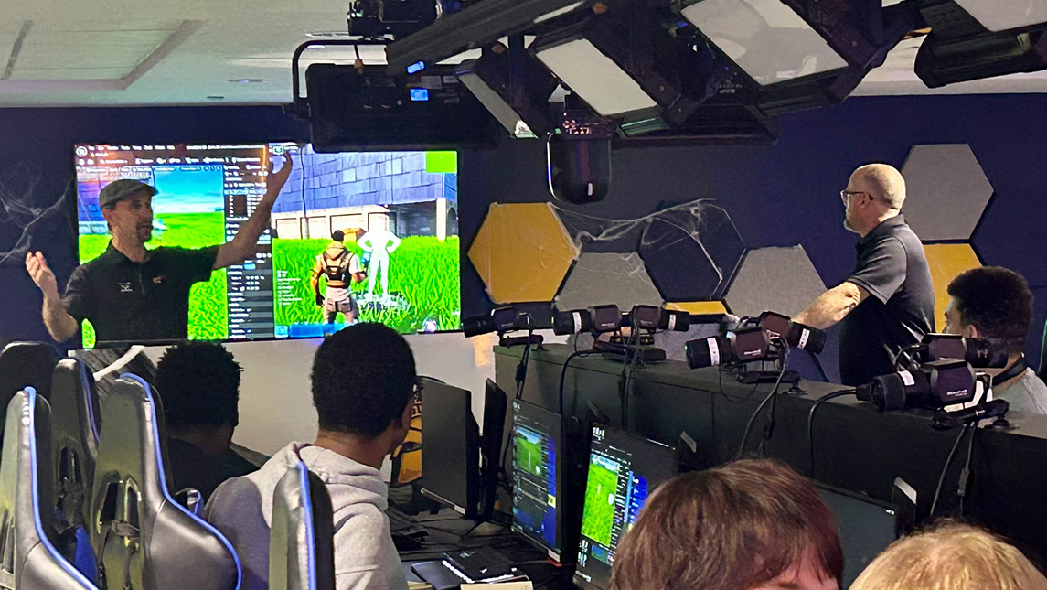 Man presents to a group of students seated at monitors in the esports arena in front of a screen with a Fortnite scene displayed.