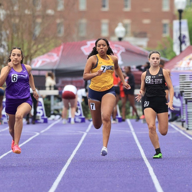 UNCG women's track runner speeds to finish with competitors on a purple lined track.