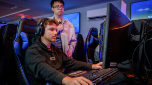 A UNCG student sits at a PC in the esports arena while another student watches.