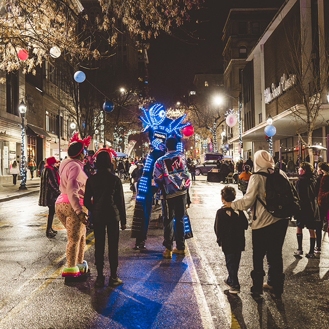 Man in stilts in a robot costume covered in blue lights walks down a downtown street interacting with people with holiday lights decorating storefronts in the background.