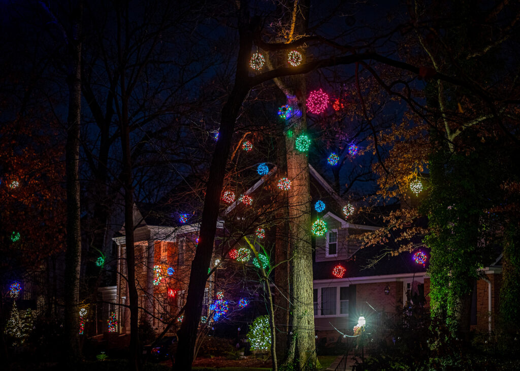Lighted holiday balls hang in trees in front of stately homes.