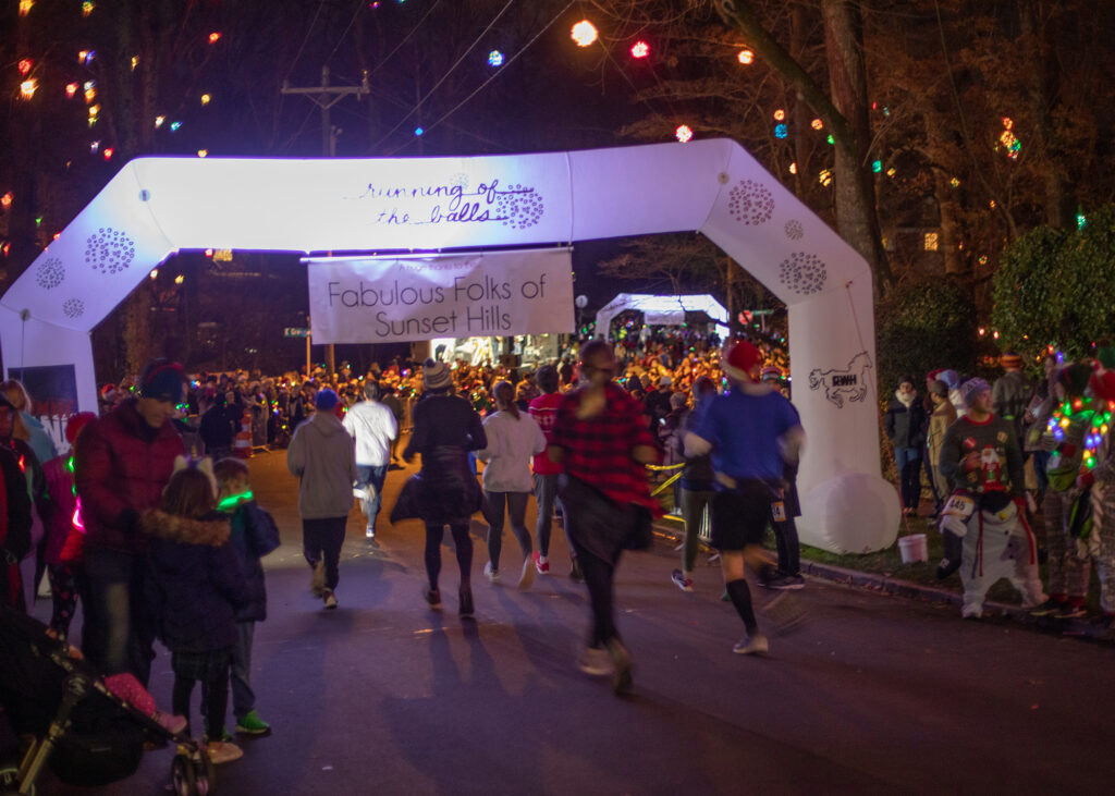 Runners in silly holiday attire walk under an arch that reads "Running of the Balls" with people lining the street on either side and lighted balls in the trees illuminate a dark sky.