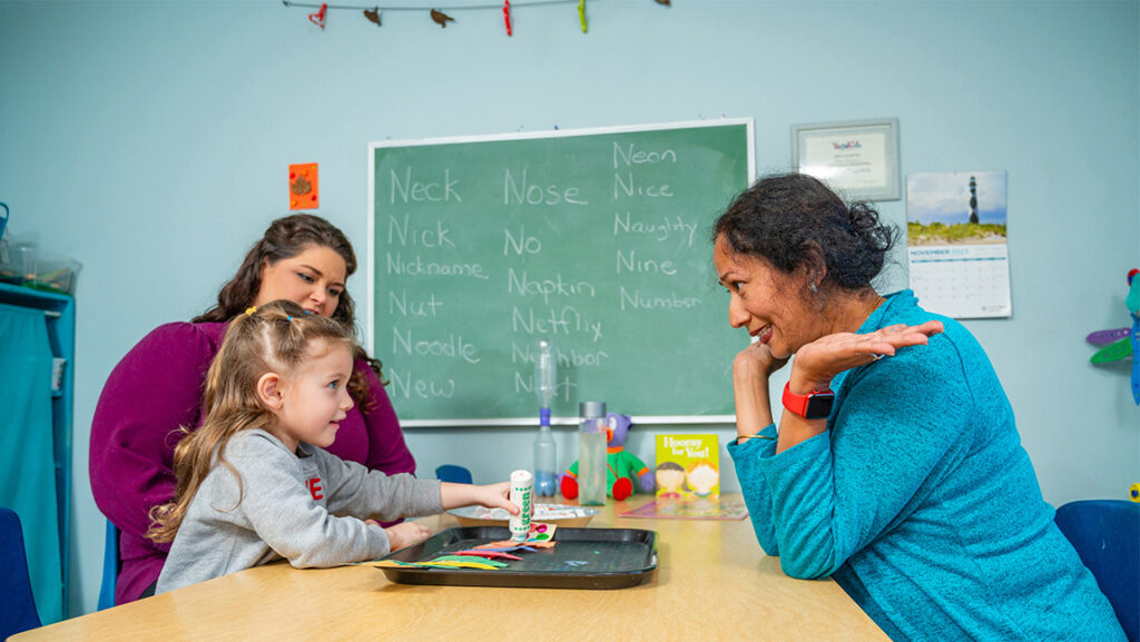 A teacher sits at a classroom table across from a child who points at pieces of paper on a tray.