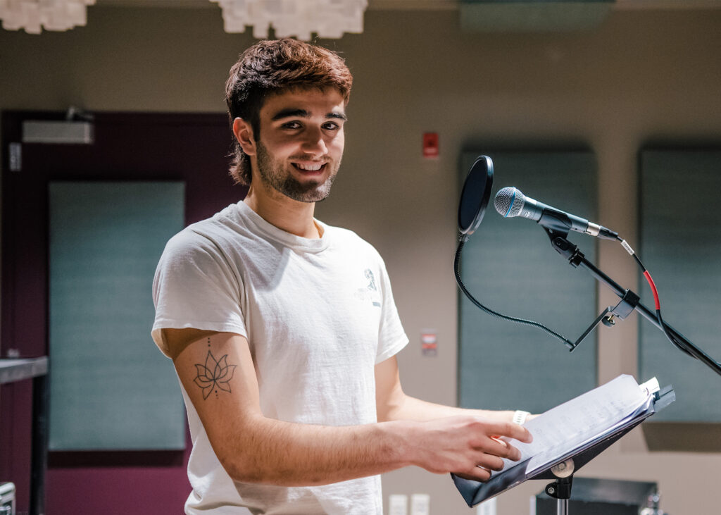 Student poses in a sound studio at a music stand with a microphone.