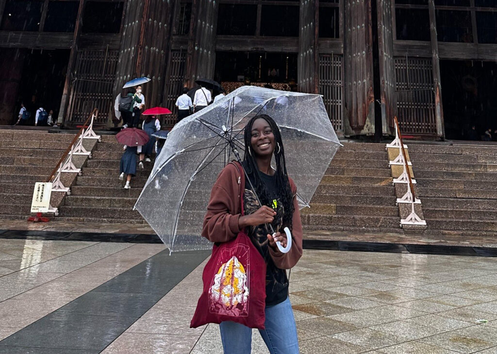 Student stands in front of a grand building with an umbrella. 