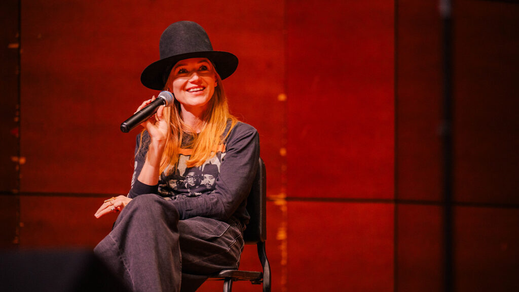 Jewel smiles with a microphone in hand while sitting on stage at the Tew Recital Hall