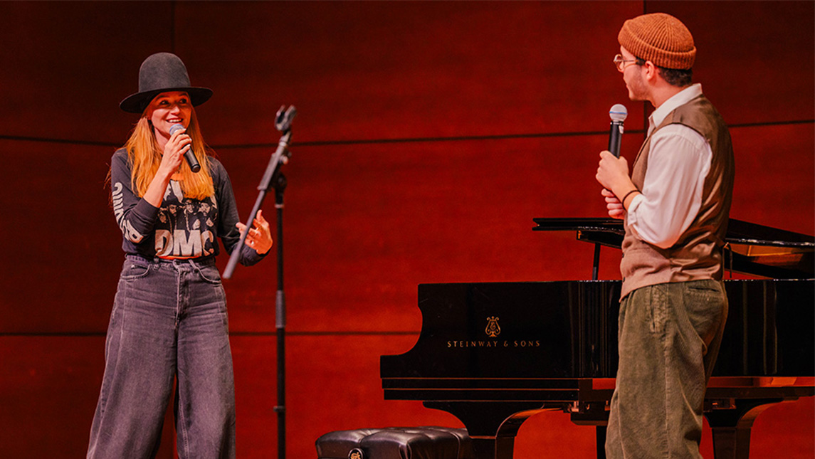 A woman (Jewel) speaks with a UNCG student on stage at the Tew Recital Hall. Both are holding microphones.
