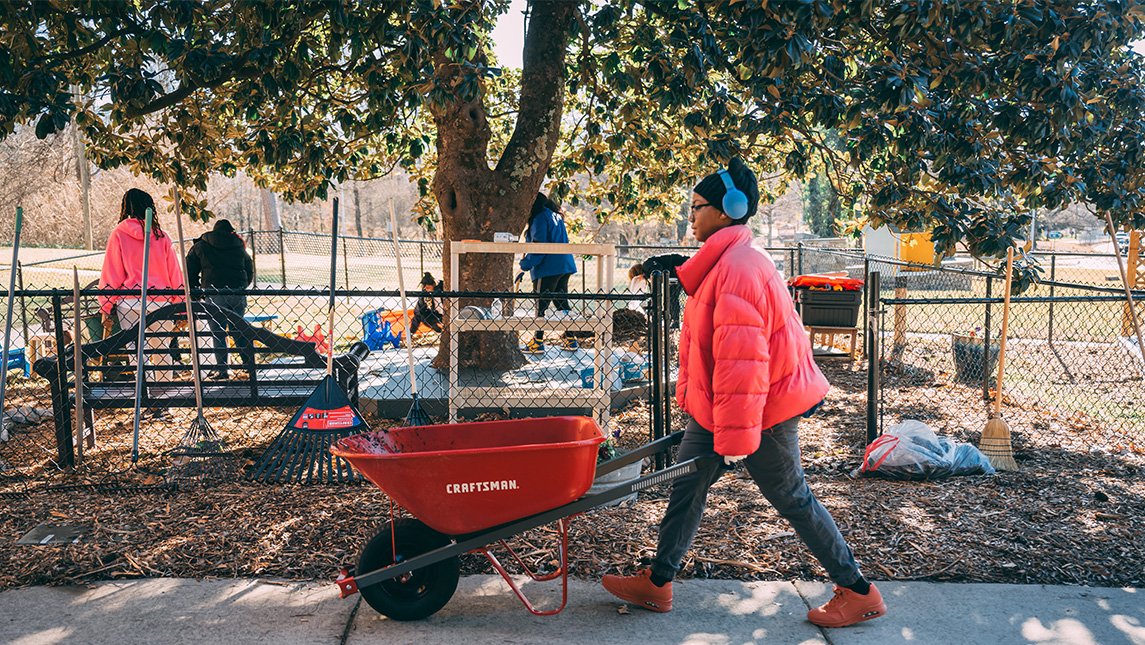 Student pushes a wheelbarrow full of mulch with others working to clean up a playground in the background. Student