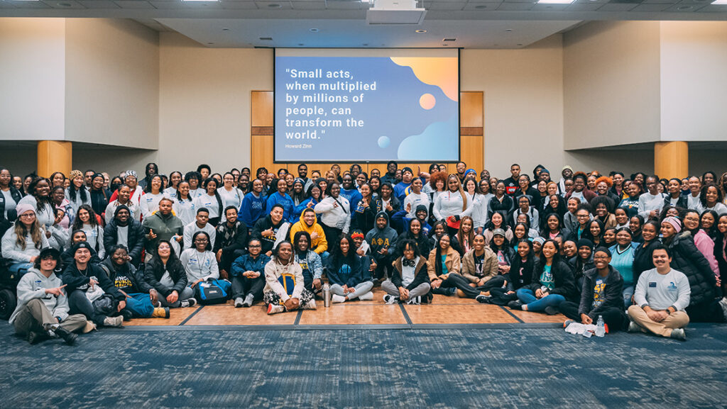 MLK Day of Service group of 200 students pose in front of a screen featuring a quote about service.