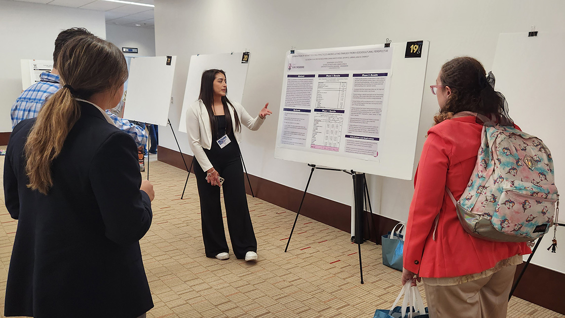 A woman stands in front of a research poster, while two other individuals listen to her speak.