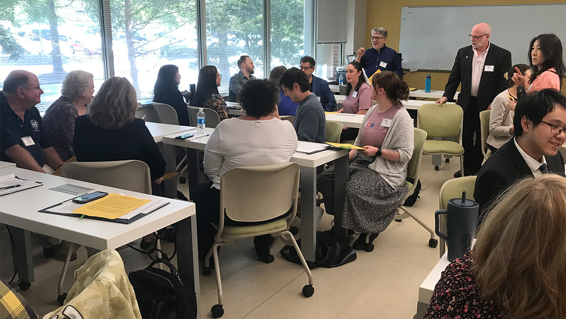 A crowd of people sit around a table and talk during a conference at UNCG.