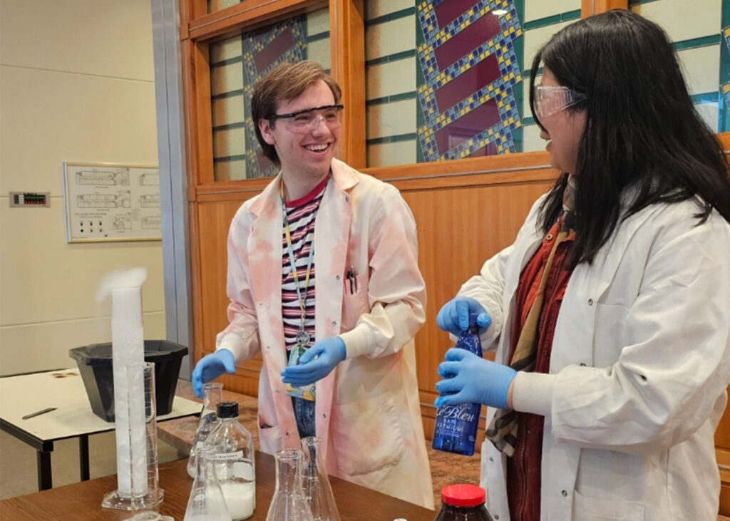 Student in a lab coat smiles at another while they do a chemistry experiment.
