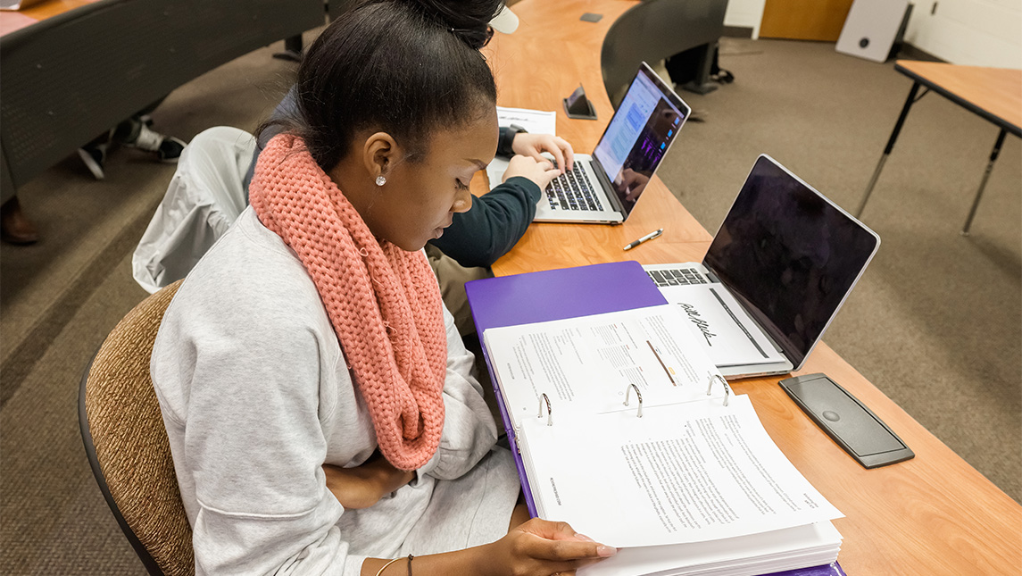 A UNCG student in a classroom turns pages in a binder beside her laptop.