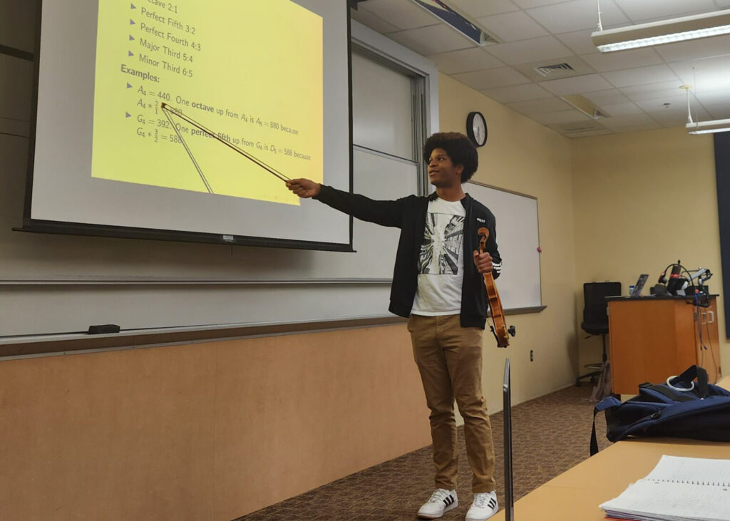 Student holds a violin in one hand as he points to a projection screen in front of a classroom. 