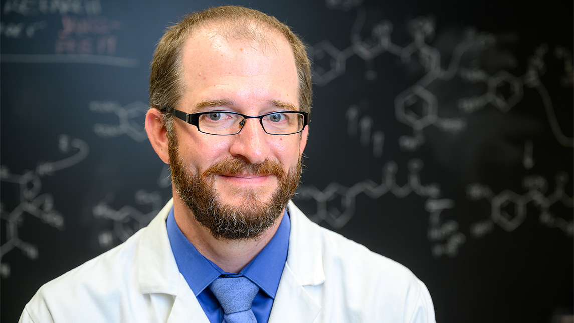 UNCG Dr. Mitch Croatt in a lab coat in front of a blackboard.