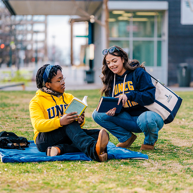 Student sits on a blanket in a park with a book and another squats to show her what she's reading. 