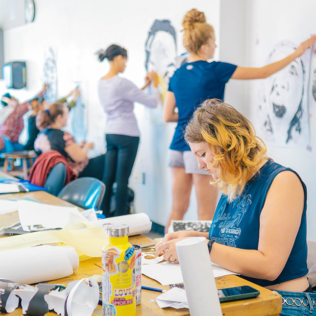 Art students in Gatewood studio working on paintings at tables with art supplies and others working on sketches hanging on the walls.  