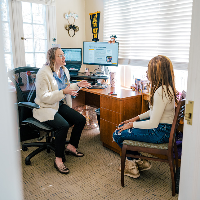 A peek into an office decorated with UNCG penants with a student talking to an advisor.