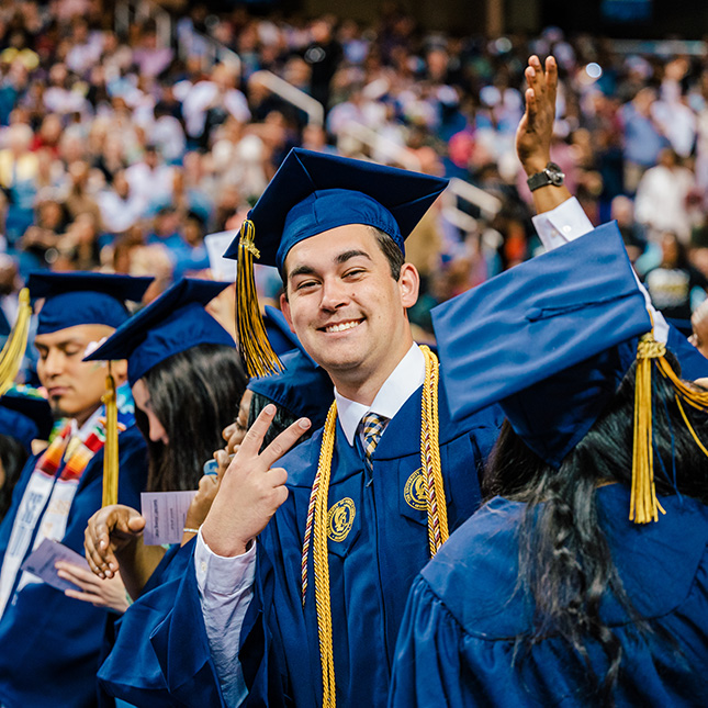 Graduate in a crowd of his peers at commencement smiles broadly and gives a peace sign to the camera.