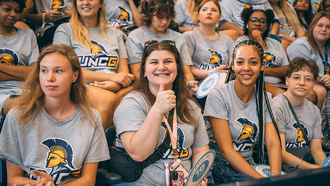 Group of students at orientation all dressed in UNCG t-shirts. The central person smiles and gives a thumbs up.