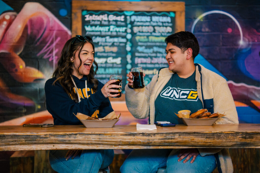 2 students clink glasses as they enjoy a bite to eat at the bar of a restaurant with a chalk board menu behind them.