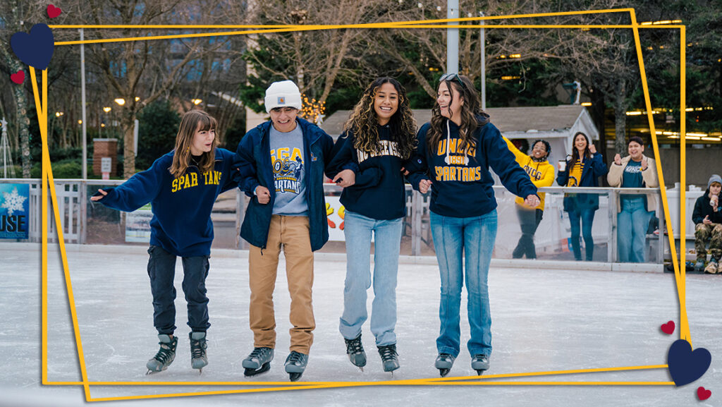 4 students in UNCG gear ice skate with their arms linked together. The shot has a graphic frame with hearts around it.