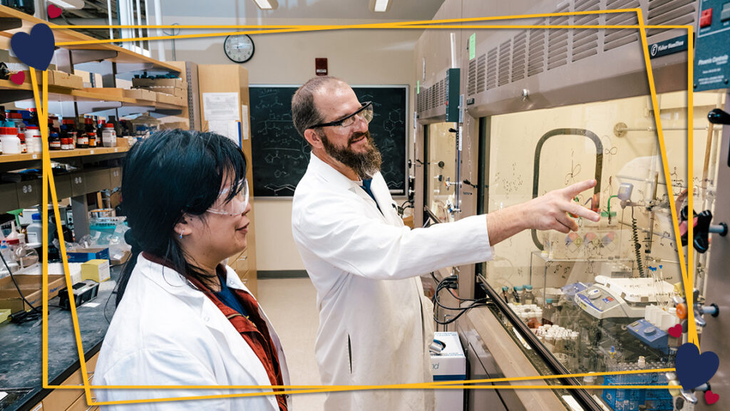 Professor and graduate student in lab coats work in a chemistry lab. Gold frame and heart graphics have been added to the photo.