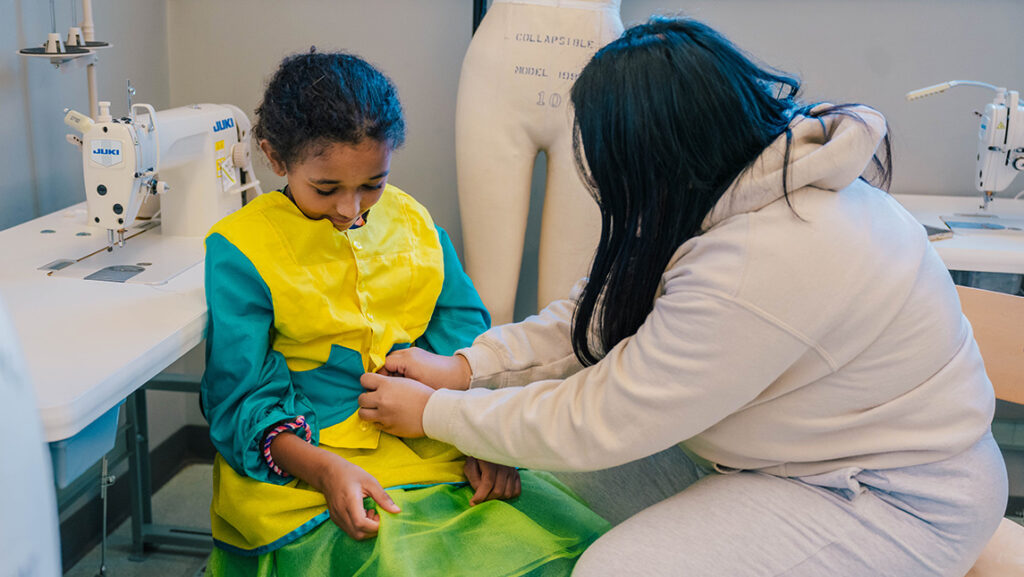 UNCG consumer, apparel, and retail studies student Lucy Buonkrong checks the fitting of a dress for Oak View Elementary student Haveah Cooper.