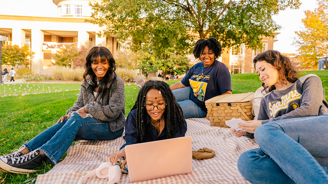 Four college students laugh together on a blanket on campus lawn with books and laptops.
