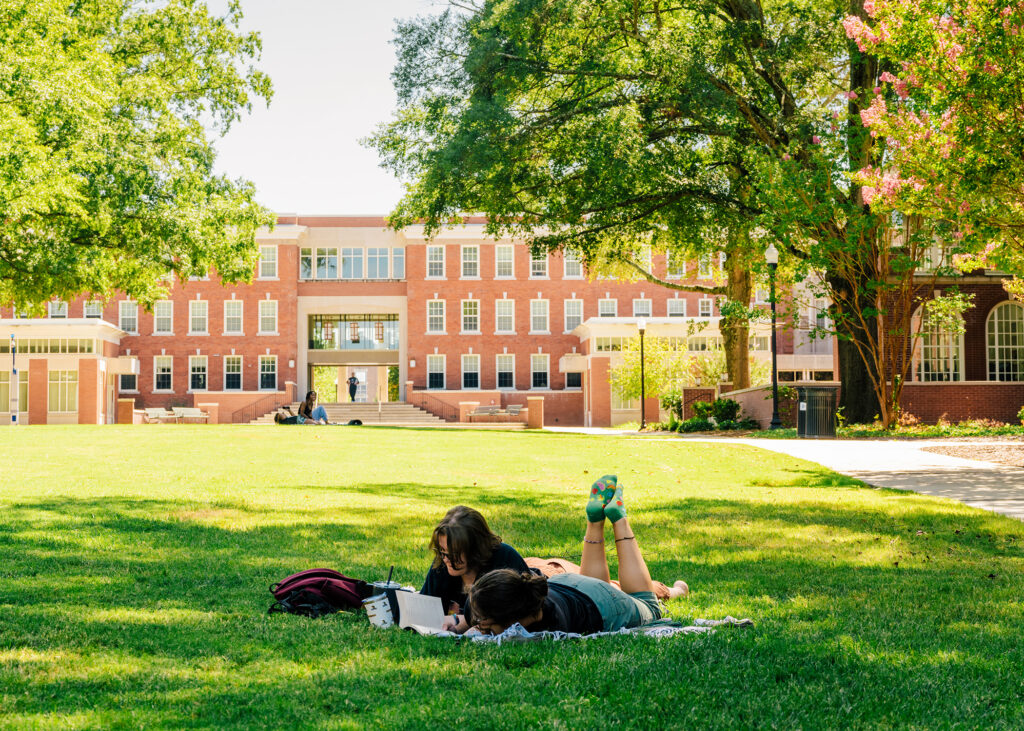 Wide shot of campus quad with a dorm building and trees in the background and a student laying on a blanket reading in the foreground.