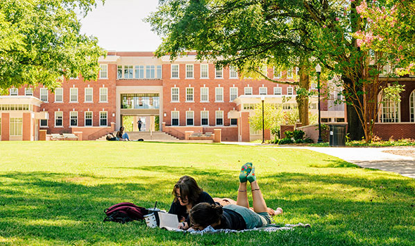 Student reads on a blanket in UNCG's quad lawn with a residence hall in the background. 