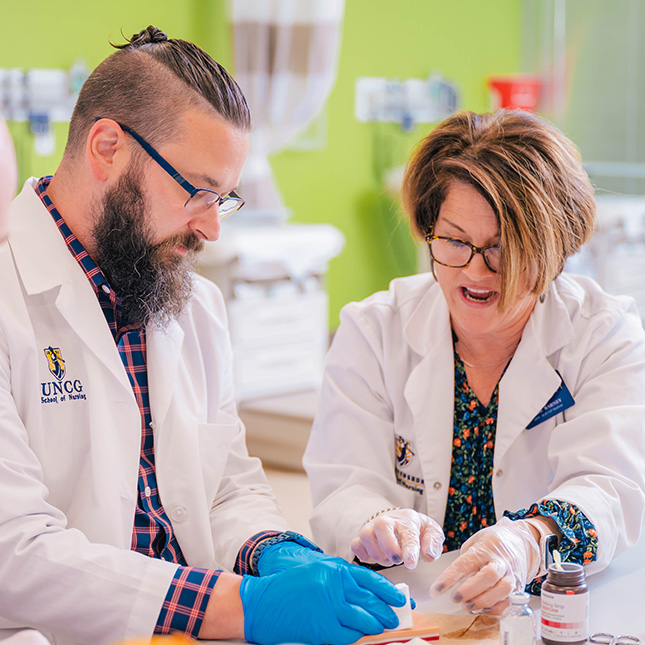 A teacher and student in white nursing lab coats practice suturing.
